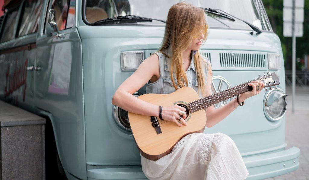 Hippie girl plays on a mini acoustic guitar leaning against a classic Volkswagen