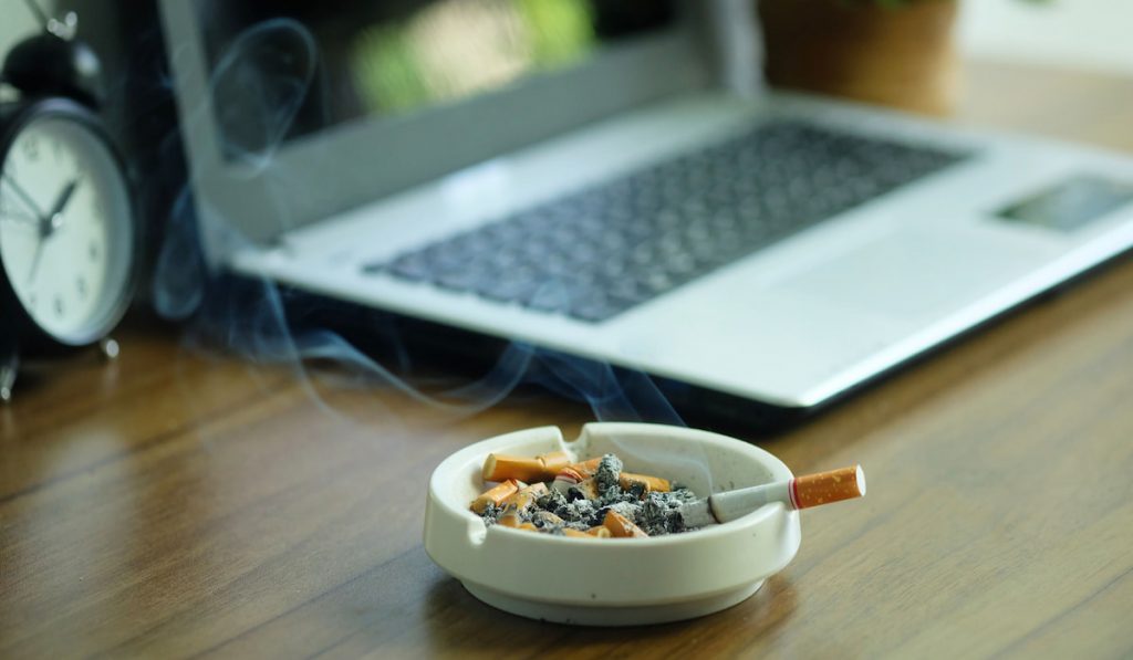 Many cigarette in white ashtray on the desk with laptop computer.