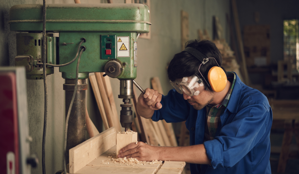 Asian young carpenter using drill press to made hole in wooden plank

