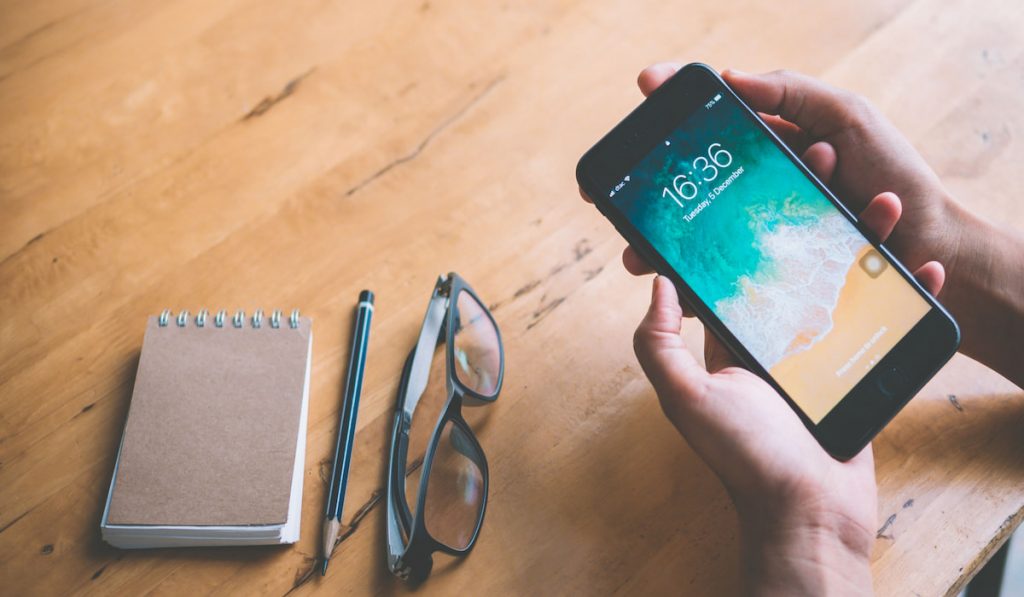 iPhone showing lock screen  on a hand  on a table with notebook, eyeglasses and pencil