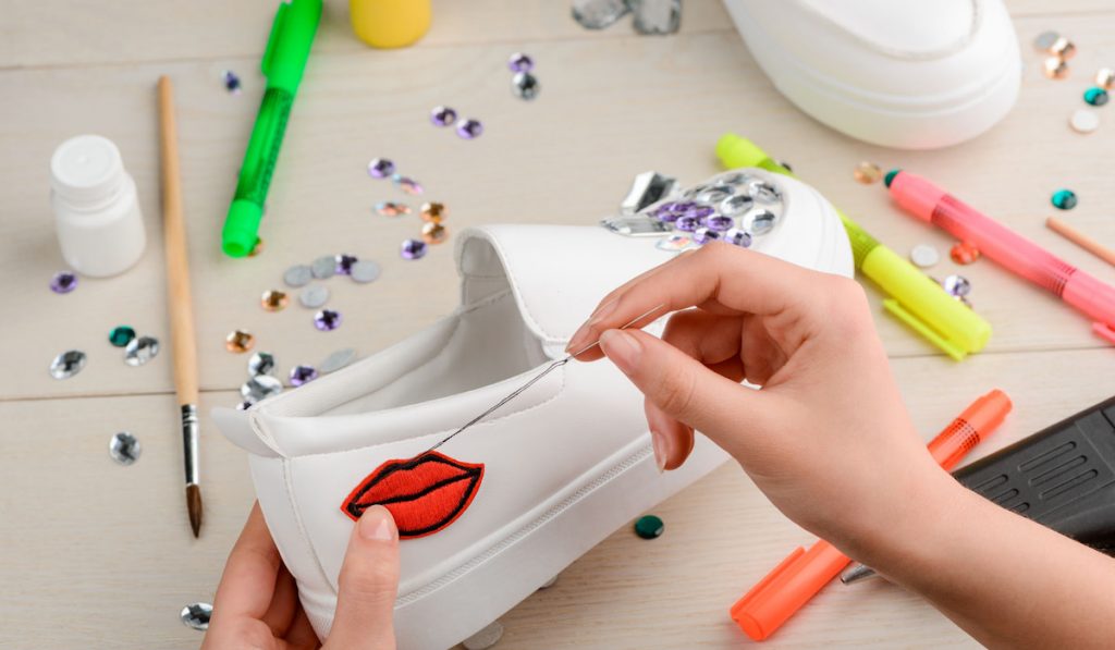 Designer sewing embroidered lips badge to a white sneaker. Rhinestones, pens and paints on a craft table.
