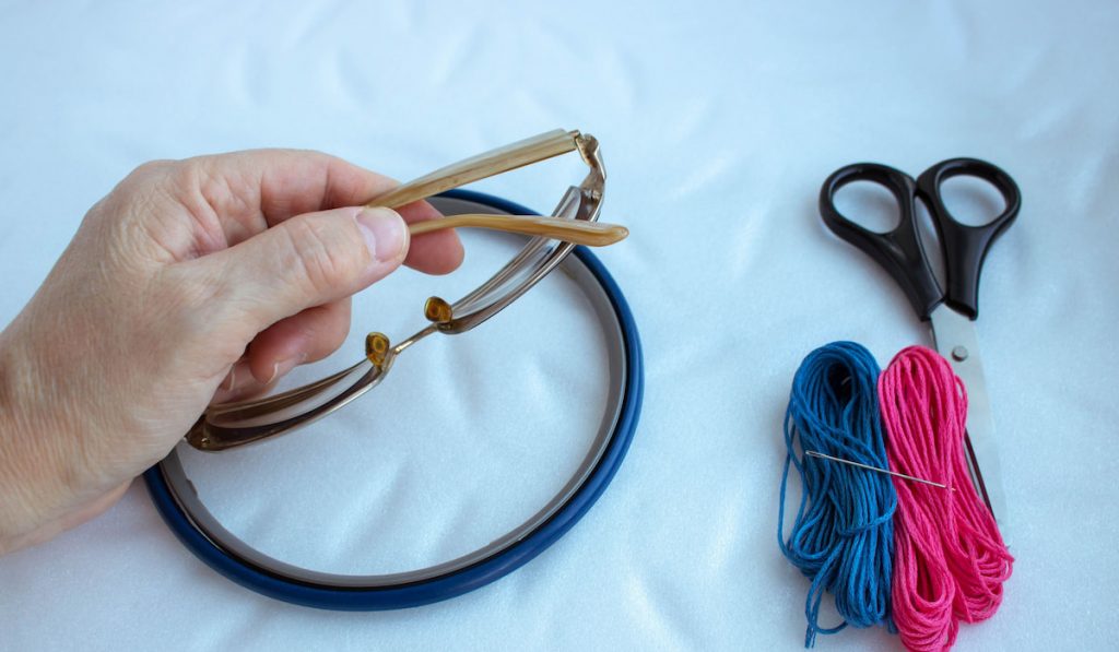 Emobroidery frame fabric needle scissors and a hand holding eyeglasses