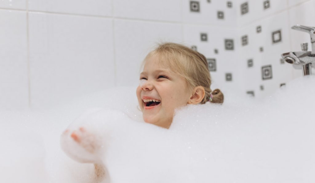 Happy Little girl baths in a bath and plays with soapy foam, enjoying bubble bath